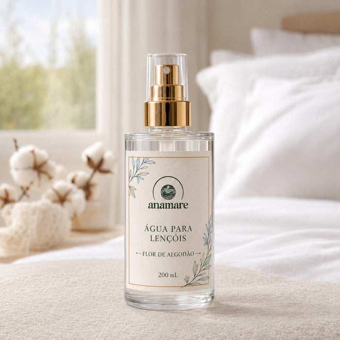 Água de Lençóis Anamare Flor de Algodão, frasco de vidro de 200 mL com válvula spray dourada e rótulo cru, em quarto claro com roupa de cama branca e ramos de algodão