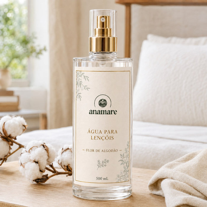 Água de Lençóis Anamare Flor de Algodão, frasco de vidro de 500 mL com válvula spray dourada e rótulo cru, em quarto claro com roupa de cama branca e ramos de algodão