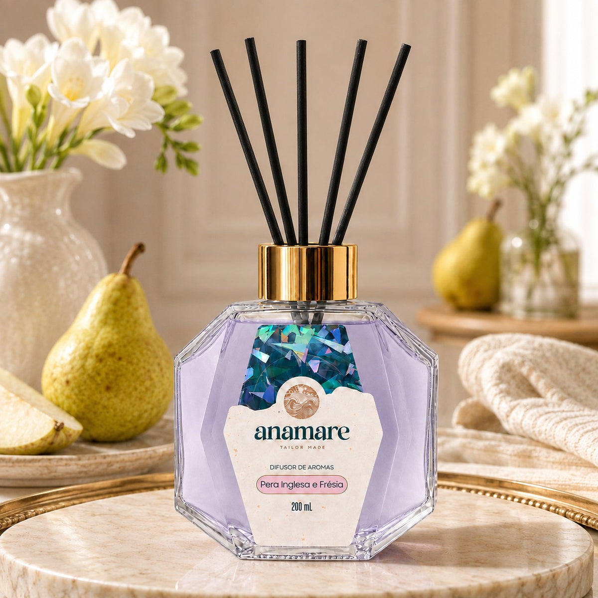 Difusor de Aromas Anamare Pera Inglesa e Frésia de 200 mL, frasco octogonal facetado de vidro com tampa dourada e varetas pretas, em bandeja dourada sobre mármore, com peras partidas em prato e frésias brancas em vaso, em luz de janela