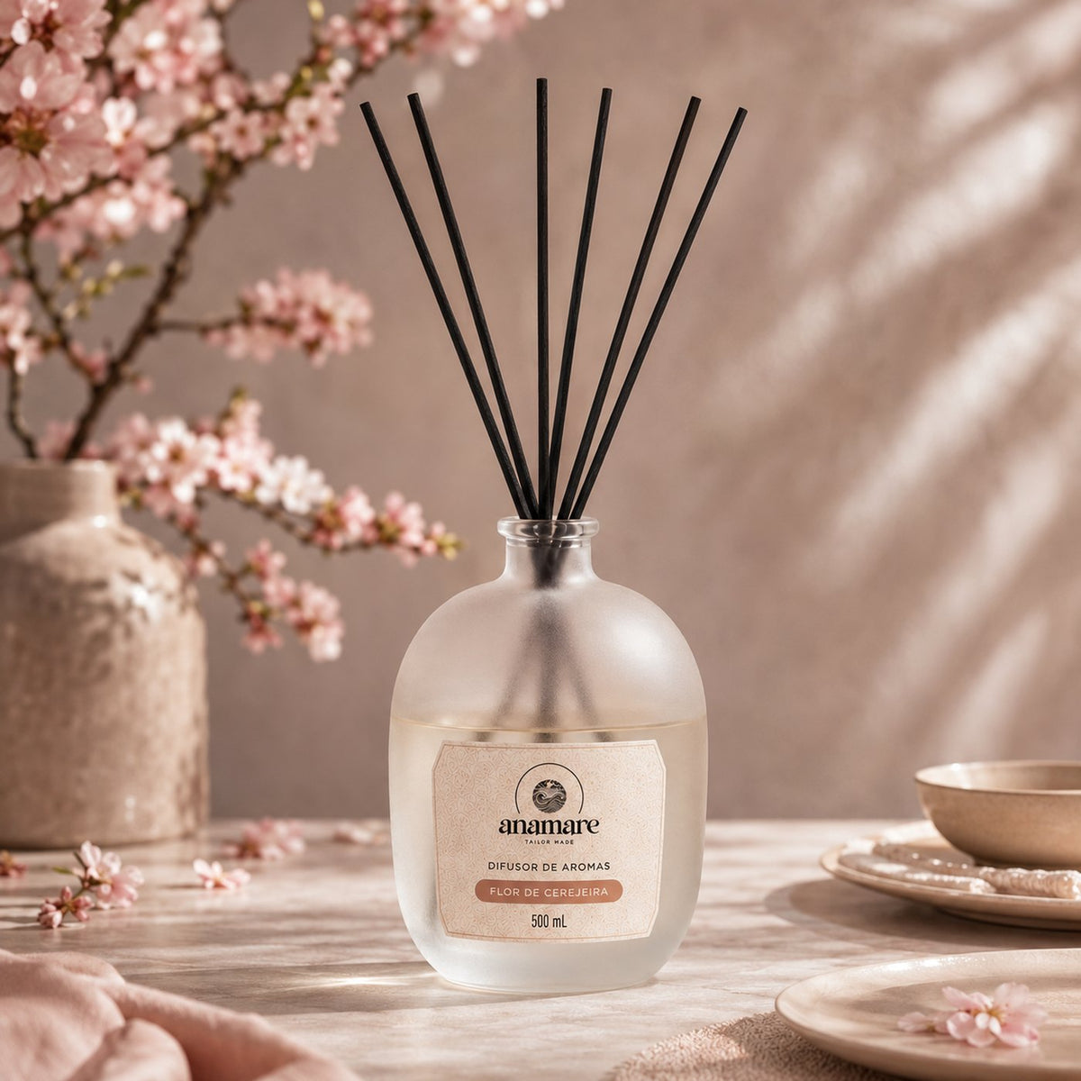 Difusor de Aromas Anamare Flor de Cerejeira de 500 mL, frasco de vidro fosco de ombro alto com gargalo prateado e varetas pretas, em mármore claro, diante de galho de cerejeira em flor em vaso de cerâmica, com pratos e pétalas soltas