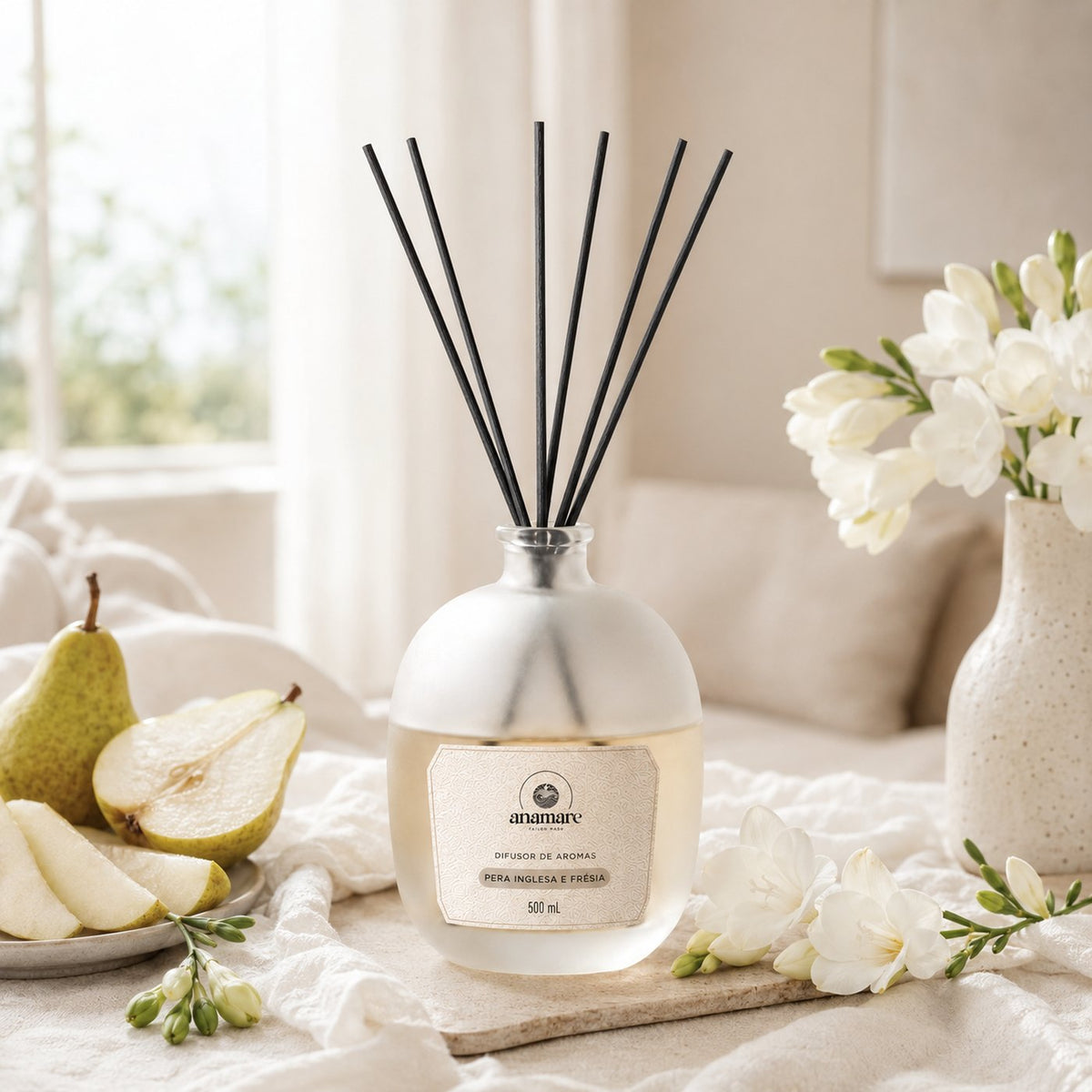 Difusor de Aromas Anamare Pera Inglesa e Frésia de 500 mL, frasco de vidro fosco de ombro alto com gargalo prateado e varetas pretas, em bandeja de pedra clara junto à janela, com peras partidas e frésias brancas em vaso de cerâmica, em luz da manhã