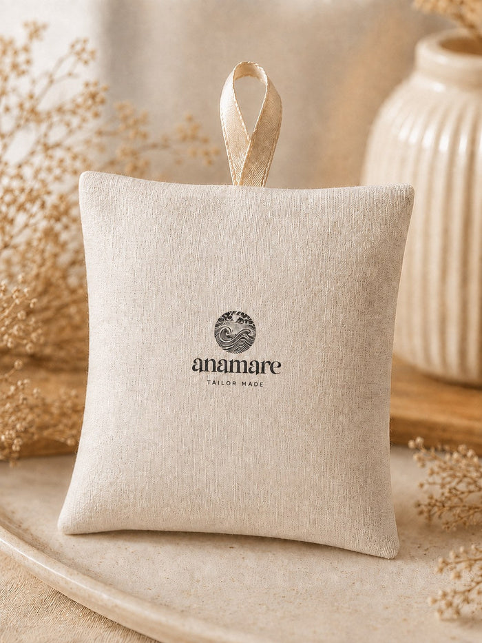 Sachê Aromático Anamare em linho cru com o selo da marca impresso e fita de cetim para pendurar, sobre prato de cerâmica bege, com flores secas e vaso canelado ao fundo em luz suave