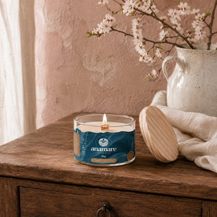 Vela Aromática Flor de Cerejeira Moyen 90 g acesa, pote de vidro com pavio de madeira, em mesa de madeira, com ramo de cerejeira em flor em jarra branca, parede rosada e tampa de madeira ao lado