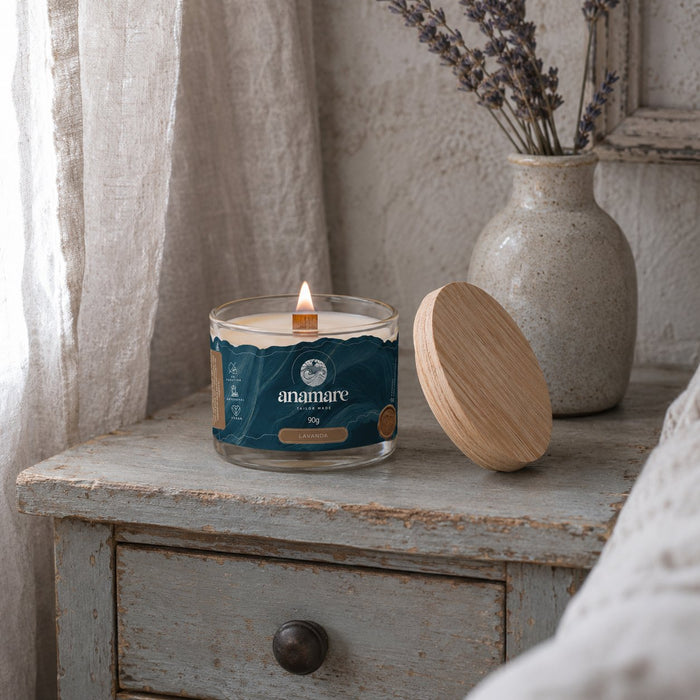 Vela Aromática Lavanda Moyen 90 g acesa, pote de vidro com pavio de madeira, em mesa de cabeceira envelhecida, com ramos de lavanda em vaso de cerâmica, cortina de linho e tampa de madeira ao lado
