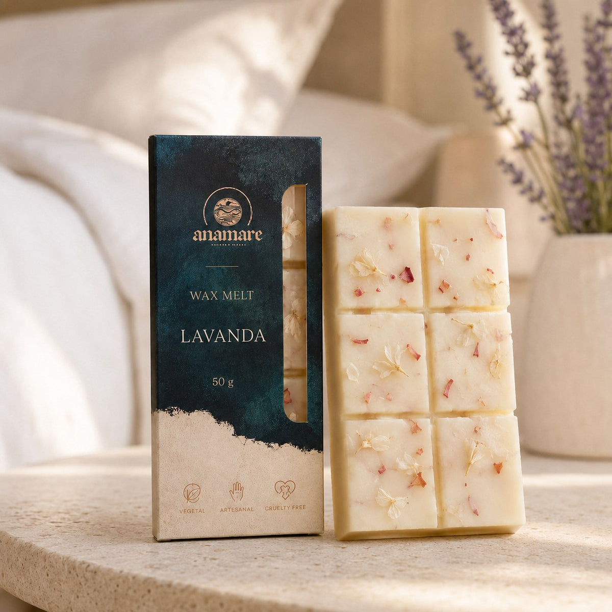 Wax Melt Anamare Lavanda de 50 g, barra de cera vegetal com pétalas secas ao lado da caixa azul-petróleo com janela, em cabeceira de cama com roupa de cama branca e lavandas em vaso de cerâmica, sobre mesa lateral de travertino
