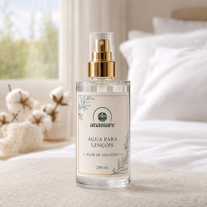 Água de Lençóis Anamare Flor de Algodão, frasco de vidro de 200 mL com válvula spray dourada e rótulo cru, em quarto claro com roupa de cama branca e ramos de algodão