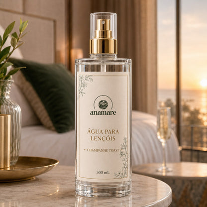 Água de Lençóis Anamare Champanhe Toast, frasco de vidro de 500 mL com válvula spray dourada e rótulo cru, em quarto com vista para o pôr do sol e taça de champanhe