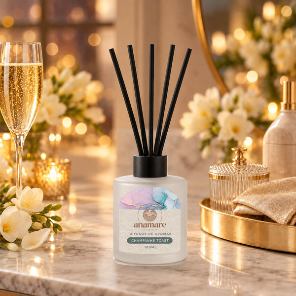 Difusor de Aromas Champanhe Toast 100 mL, frasco de vidro fosco com varetas pretas, em bancada de mármore com taça de champanhe e velas acesas ao fundo