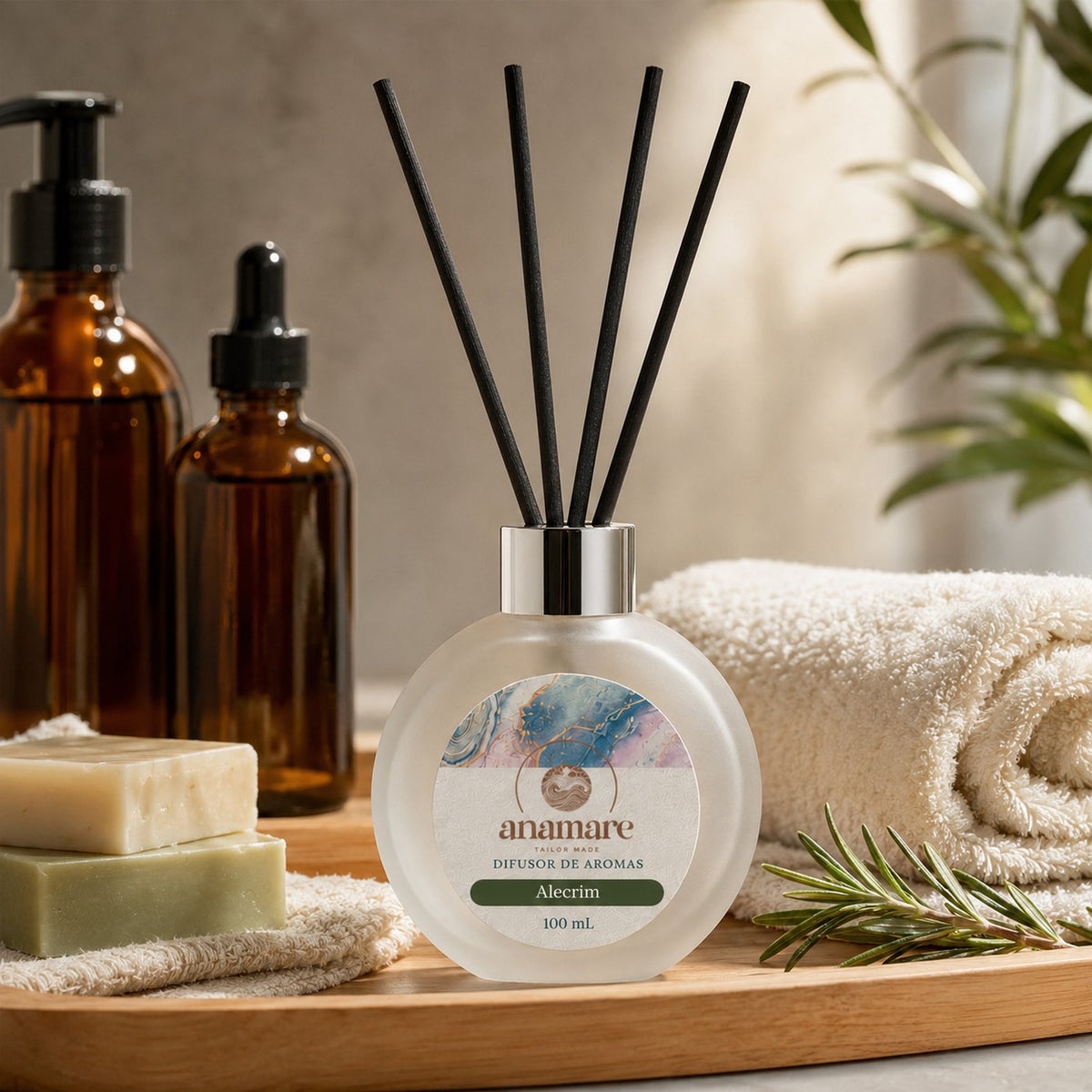 Difusor de Aromas Anamare Alecrim de 100 mL, frasco redondo de vidro fosco com tampa cromada e varetas pretas, em tábua de madeira ao lado de toalhas felpudas brancas e um ramo de alecrim fresco