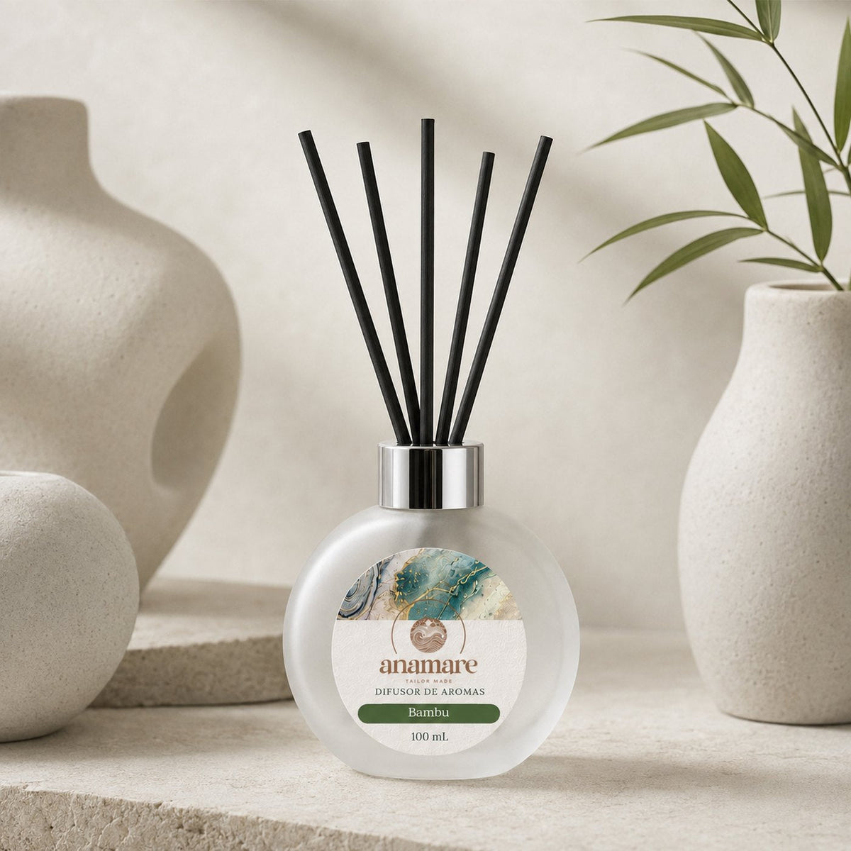 Difusor de Aromas Anamare Bambu de 100 mL, frasco redondo de vidro fosco com tampa cromada e varetas pretas, sobre superfície clara ao lado de um bloco de pedra, em luz difusa e fundo neutro