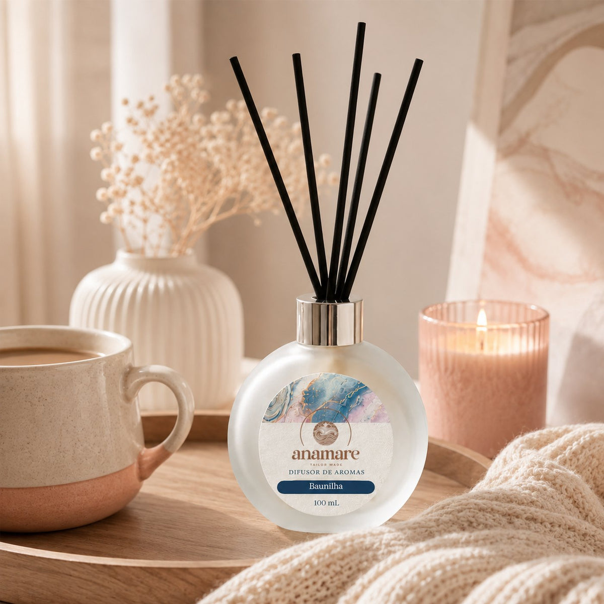 Difusor de Aromas Anamare Baunilha de 100 mL, frasco redondo de vidro fosco com tampa cromada e varetas pretas, em composição quente de tons âmbar, com madeira e tecido claro ao redor