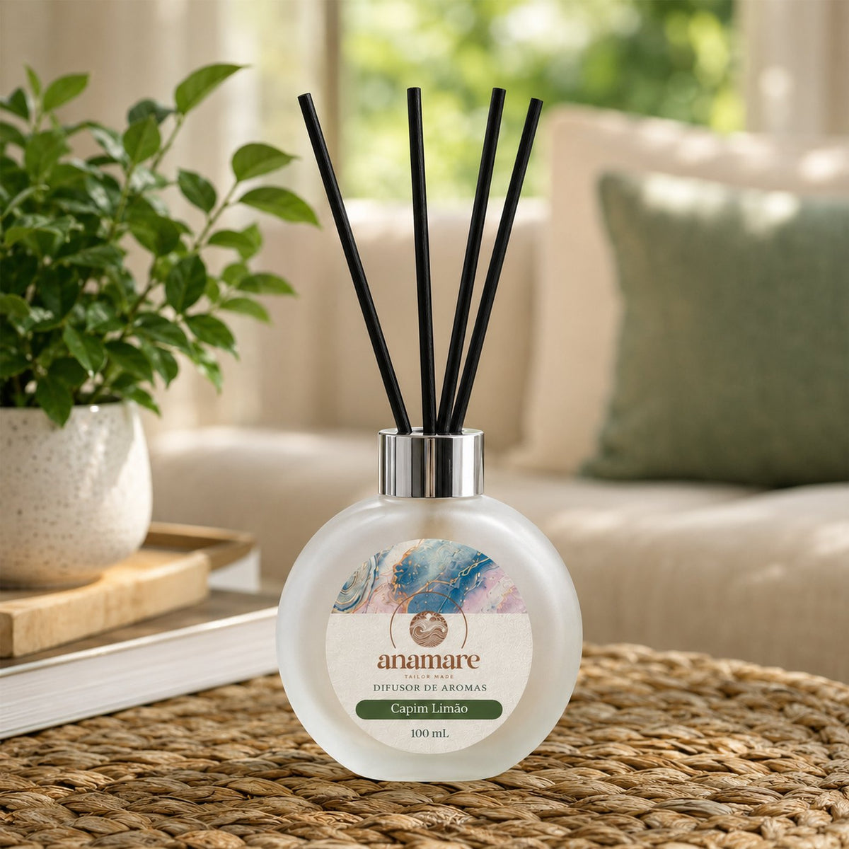 Difusor de Aromas Anamare Capim Limão de 100 mL, frasco redondo de vidro fosco com tampa cromada e varetas pretas, sobre trama de fibra natural, com planta em vaso de cerâmica, livro aberto e sofá de linho ao fundo