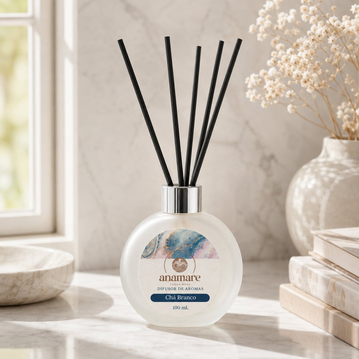 Difusor de Aromas Anamare Chá Branco de 100 mL, frasco redondo de vidro fosco com tampa cromada e varetas pretas, em peitoril de mármore ao lado da janela, com flores secas, livros empilhados e vaso de cerâmica em luz da tarde
