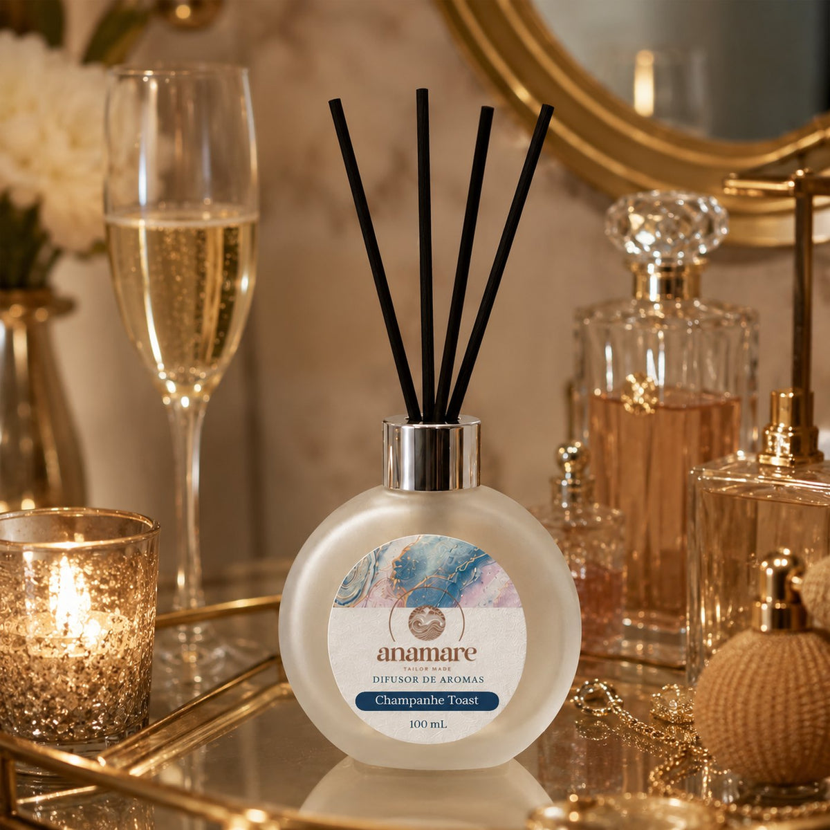 Difusor de Aromas Anamare Champanhe Toast de 100 mL, frasco redondo de vidro fosco com tampa cromada e varetas pretas, em composição elegante de tons dourados e claros, sobre superfície polida