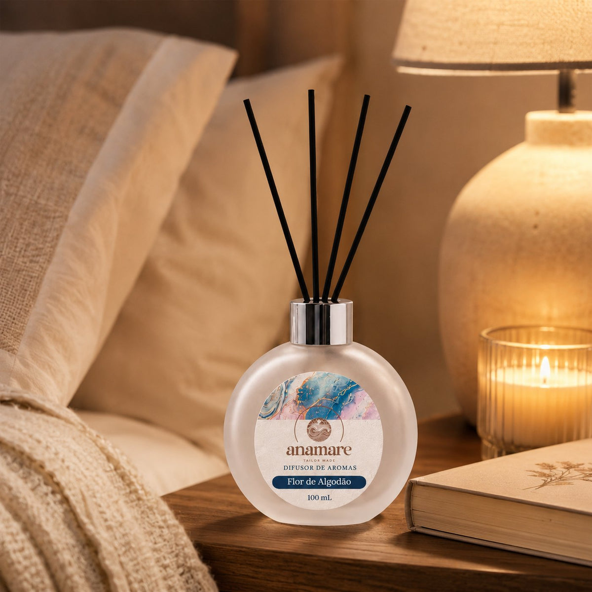 Difusor de Aromas Anamare Flor de Algodão de 100 mL, frasco redondo de vidro fosco com tampa cromada e varetas pretas, em mesa de cabeceira de madeira, com abajur aceso, vela em copo canelado e livro fechado, em luz baixa