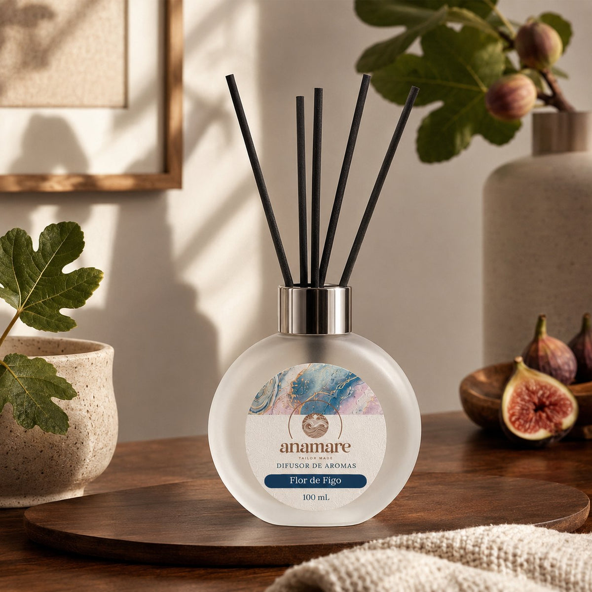 Difusor de Aromas Anamare Flor de Figo de 100 mL, frasco redondo de vidro fosco com tampa cromada e varetas pretas, em cena de tons verdes e terrosos, sobre superfície clara com fundo neutro