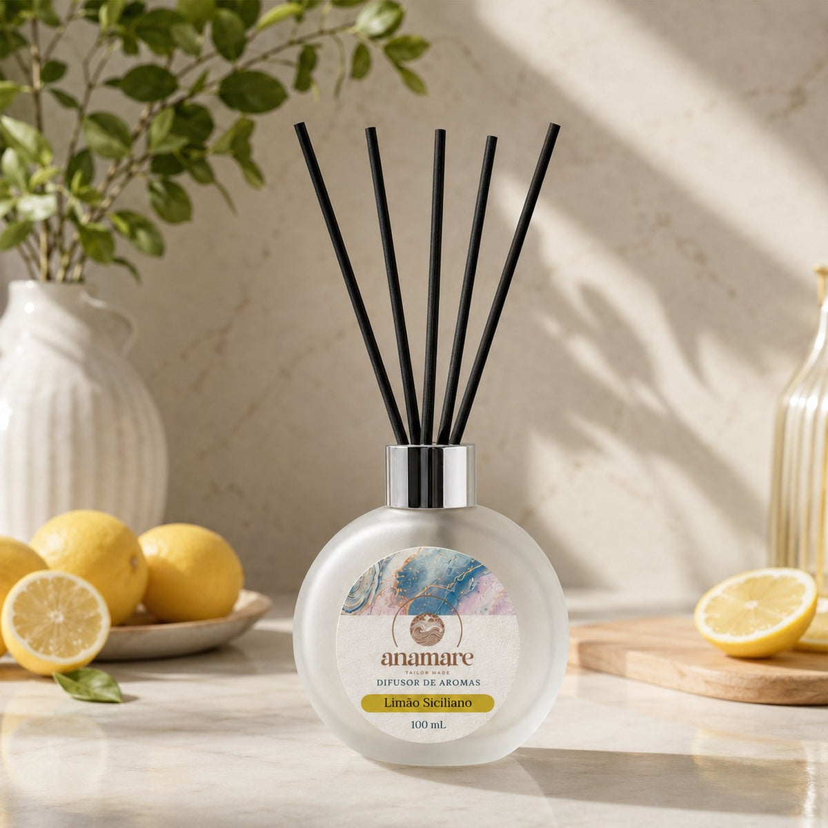 Difusor de Aromas Anamare Limão Siciliano de 100 mL, frasco redondo de vidro fosco com tampa cromada e varetas pretas, em cena clara e cítrica, sobre superfície lisa com fundo neutro