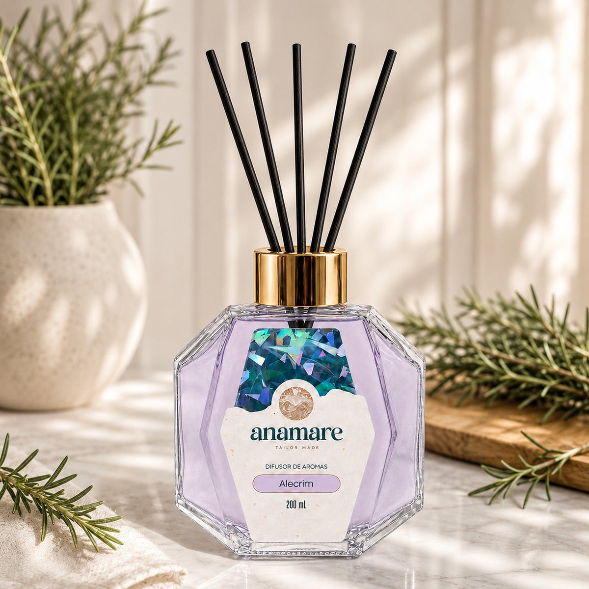 Difusor de Aromas Anamare Alecrim de 200 mL, frasco octogonal facetado de vidro com tampa dourada e varetas pretas, em bancada de mármore branco, com alecrim fresco em vaso de cerâmica, tábua de madeira e guardanapo de linho