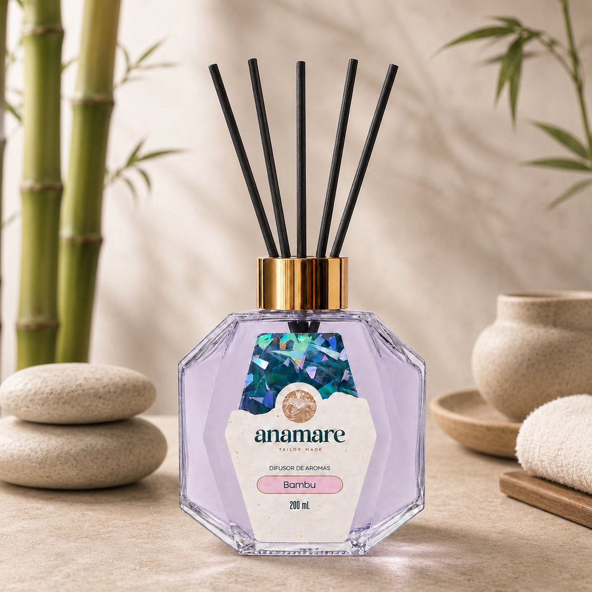 Difusor de Aromas Anamare Bambu de 200 mL, frasco octogonal facetado de vidro com tampa dourada e varetas pretas, em pedra clara de spa, com colmos de bambu, seixos empilhados, tigela de cerâmica e toalha enrolada em bandeja de madeira