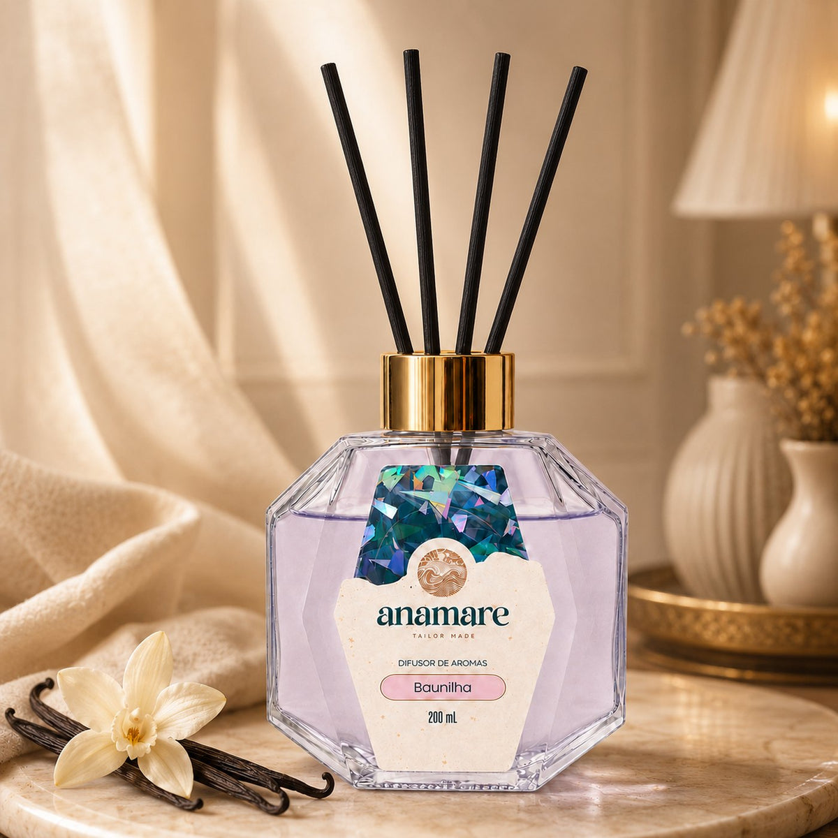 Difusor de Aromas Anamare Baunilha de 200 mL, frasco octogonal facetado de vidro com tampa dourada e varetas pretas, em bandeja dourada sobre mármore, com favas e flor de baunilha, cortina de linho e abajur aceso ao fundo