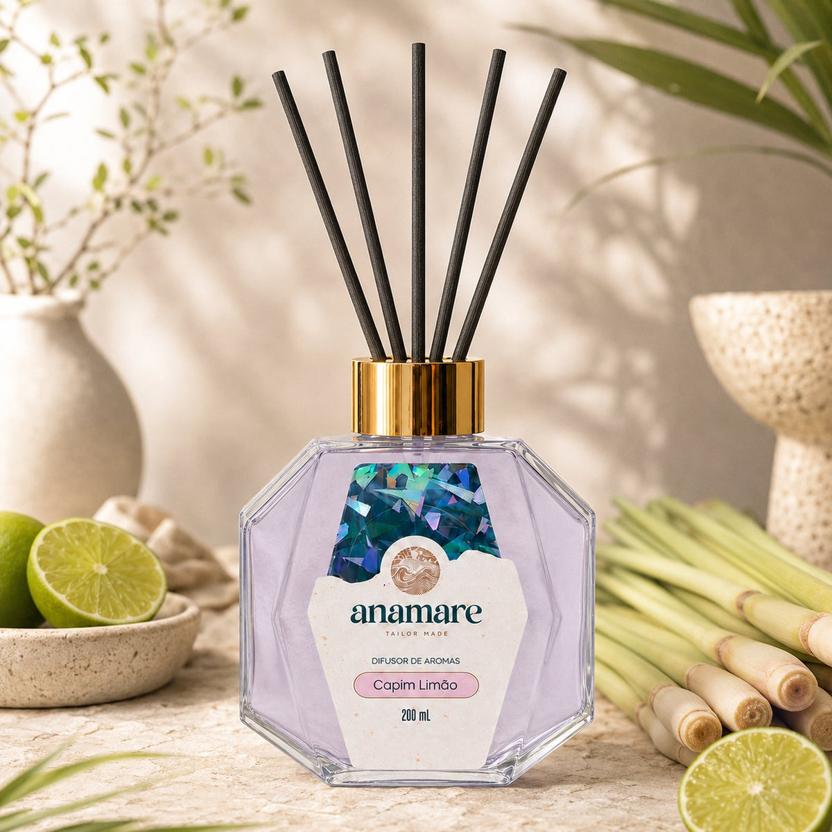 Difusor de Aromas Anamare Capim Limão de 200 mL, frasco octogonal facetado de vidro com tampa dourada e varetas pretas, em superfície de pedra clara, com talos de capim-limão, limões partidos em tigela e folhagem em vaso, em luz filtrada