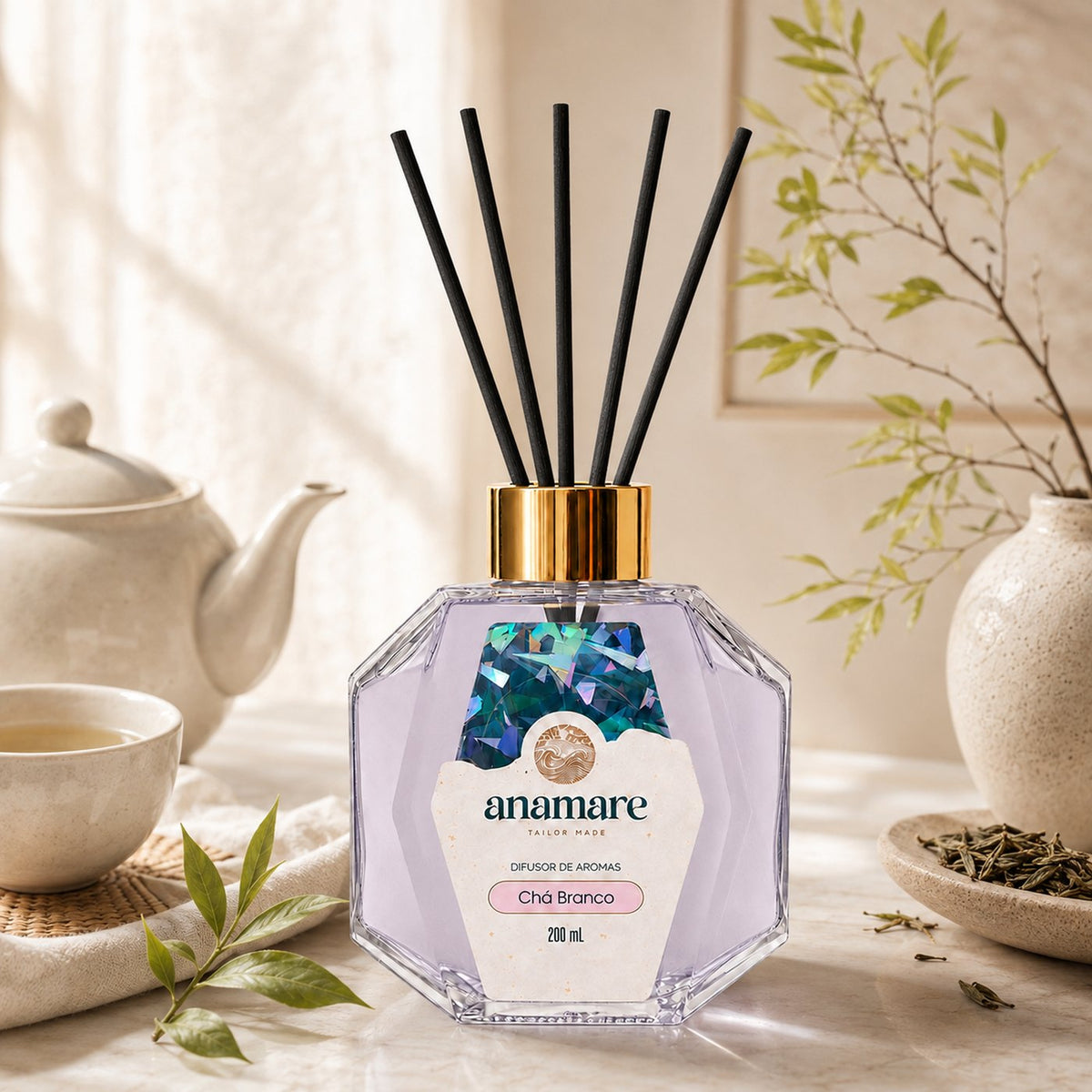Difusor de Aromas Anamare Chá Branco de 200 mL, frasco octogonal facetado de vidro com tampa dourada e varetas pretas, em bancada de mármore, com bule de porcelana, xícara de chá verde, folhas frescas e prato de folhas secas