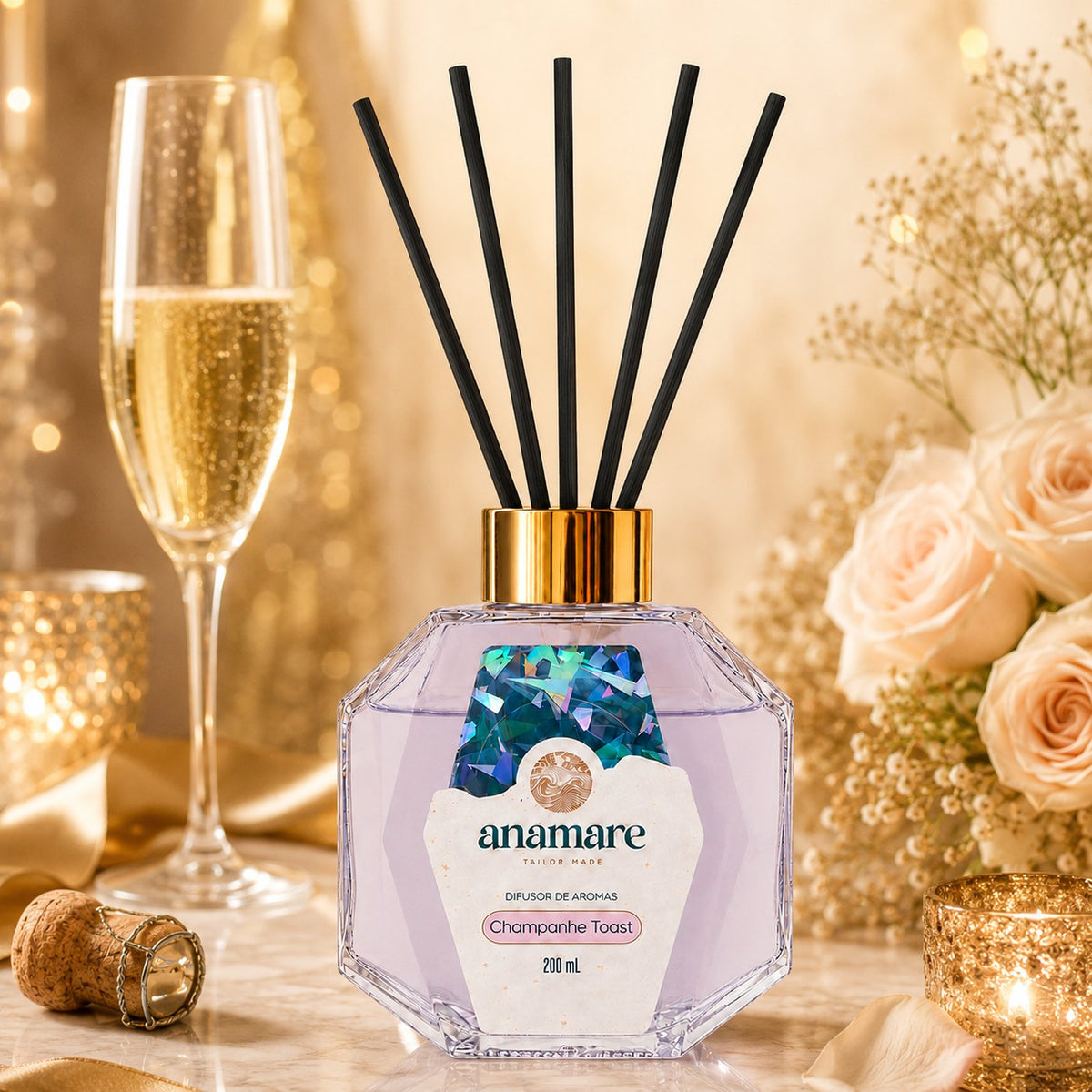 Difusor de Aromas Anamare Champanhe Toast de 200 mL, frasco octogonal facetado de vidro com tampa dourada e varetas pretas, em mármore claro, com taça de espumante, rosas champanhe, velas acesas e rolha de cortiça, em luz dourada