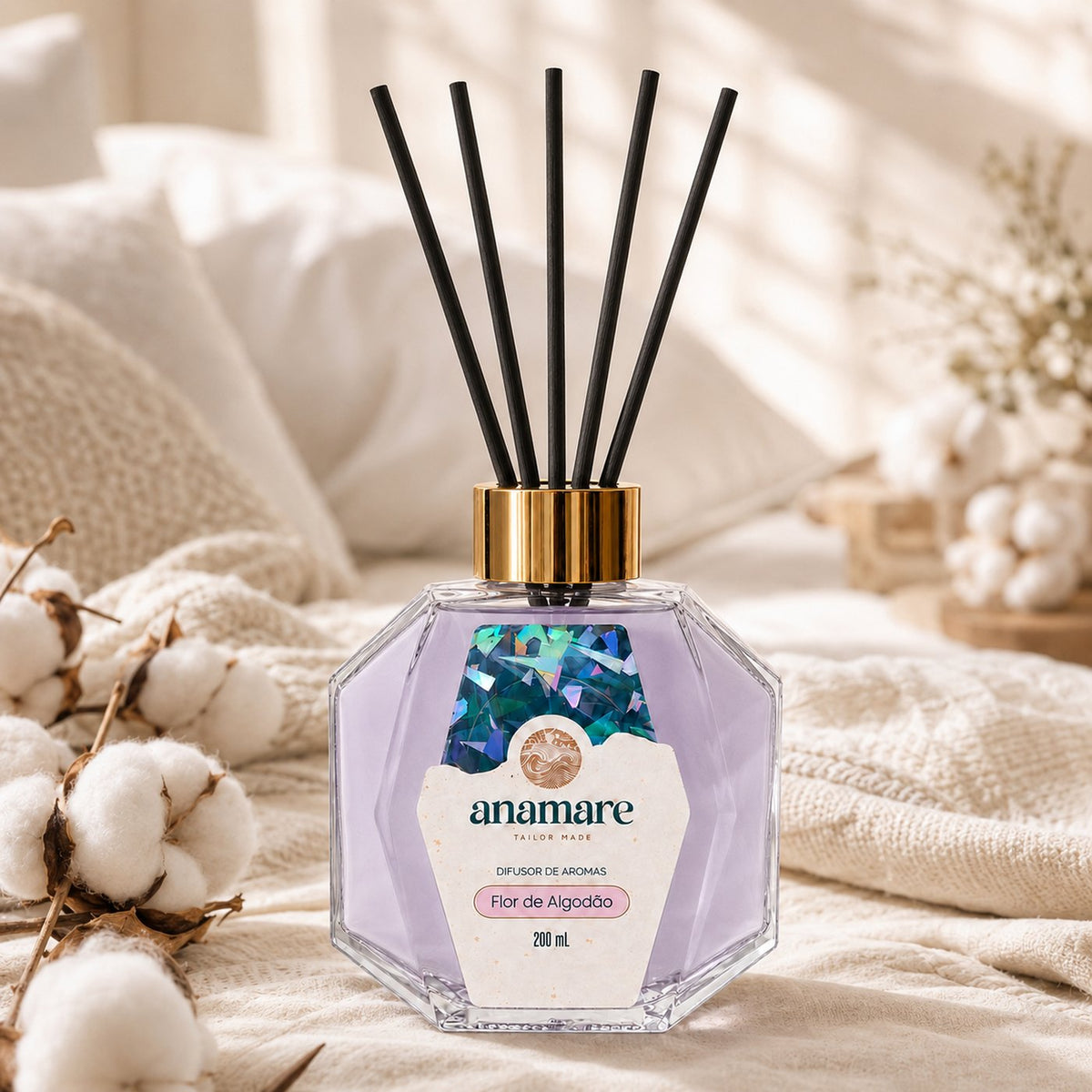 Difusor de Aromas Anamare Flor de Algodão de 200 mL, frasco octogonal facetado de vidro com tampa dourada e varetas pretas, sobre colcha de tricô creme, com ramos de algodão, almofadas brancas e luz de persiana ao fundo
