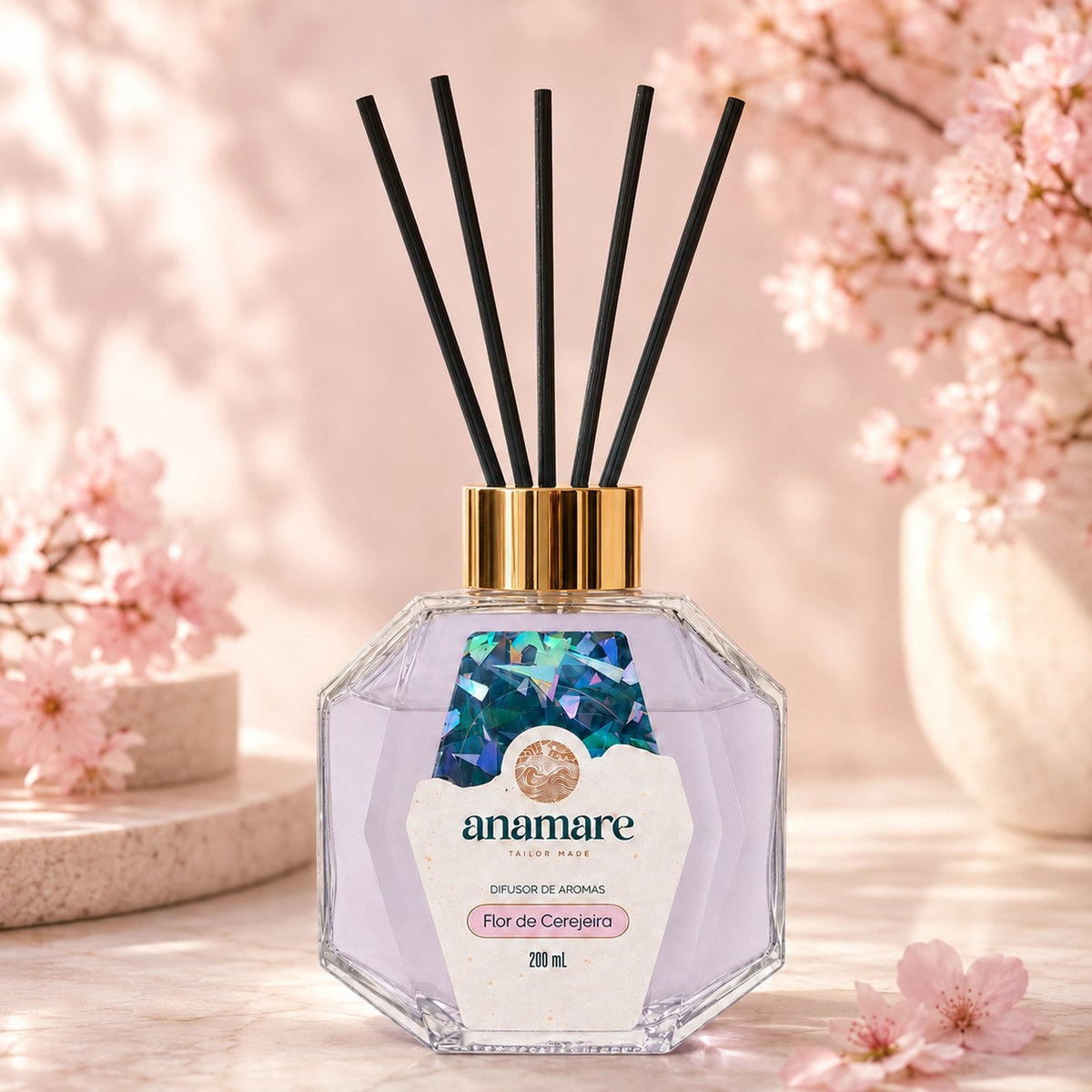 Difusor de Aromas Anamare Flor de Cerejeira de 200 mL, frasco octogonal facetado de vidro com tampa dourada e varetas pretas, em mármore rosado, diante de galhos de cerejeira em flor em vaso claro, com pódio de pedra e pétalas soltas