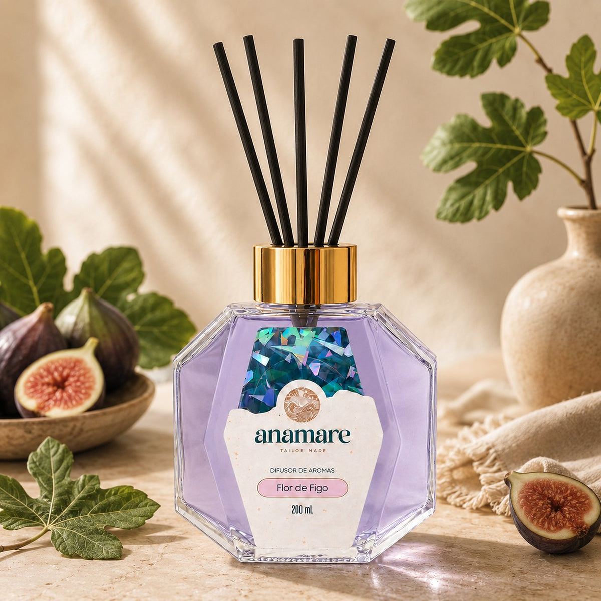 Difusor de Aromas Anamare Flor de Figo de 200 mL, frasco octogonal facetado de vidro com tampa dourada e varetas pretas, em pedra travertino, com figos partidos em tigela, folhas de figueira e jarra de cerâmica, em luz de veneziana