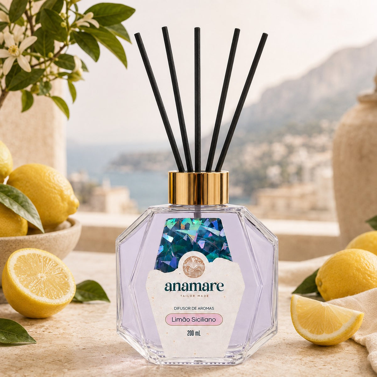 Difusor de Aromas Anamare Limão Siciliano de 200 mL, frasco octogonal facetado de vidro com tampa dourada e varetas pretas, em terraço de pedra com vista para o mar, com limões-sicilianos em tigela e galho de limoeiro em flor