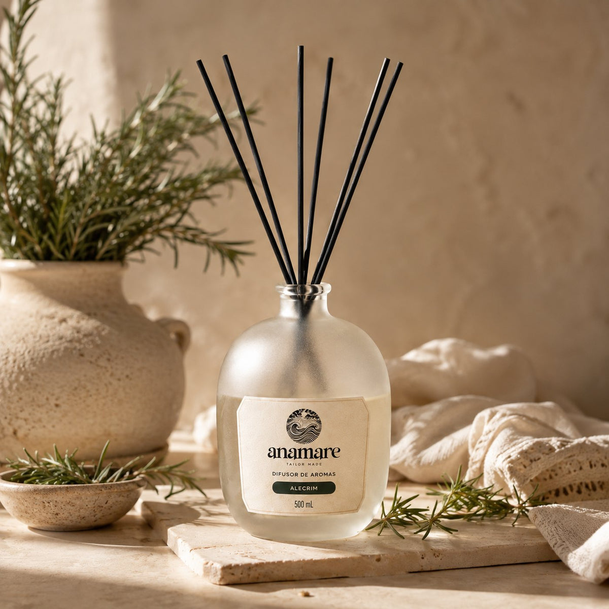 Difusor de Aromas Anamare Alecrim de 500 mL, frasco de vidro fosco de ombro alto com gargalo prateado e varetas pretas, em bandeja de madeira clara, com ramos de alecrim fresco e toalhas felpudas brancas