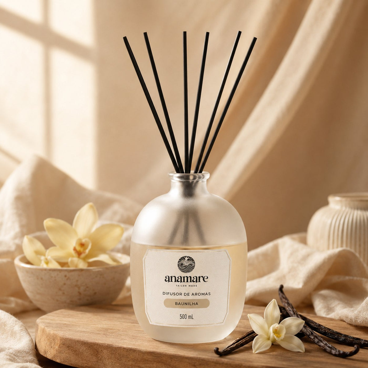 Difusor de Aromas Anamare Baunilha de 500 mL, frasco de vidro fosco de ombro alto com gargalo prateado e varetas pretas, em composição quente de tons âmbar, com madeira e tecido claro ao redor