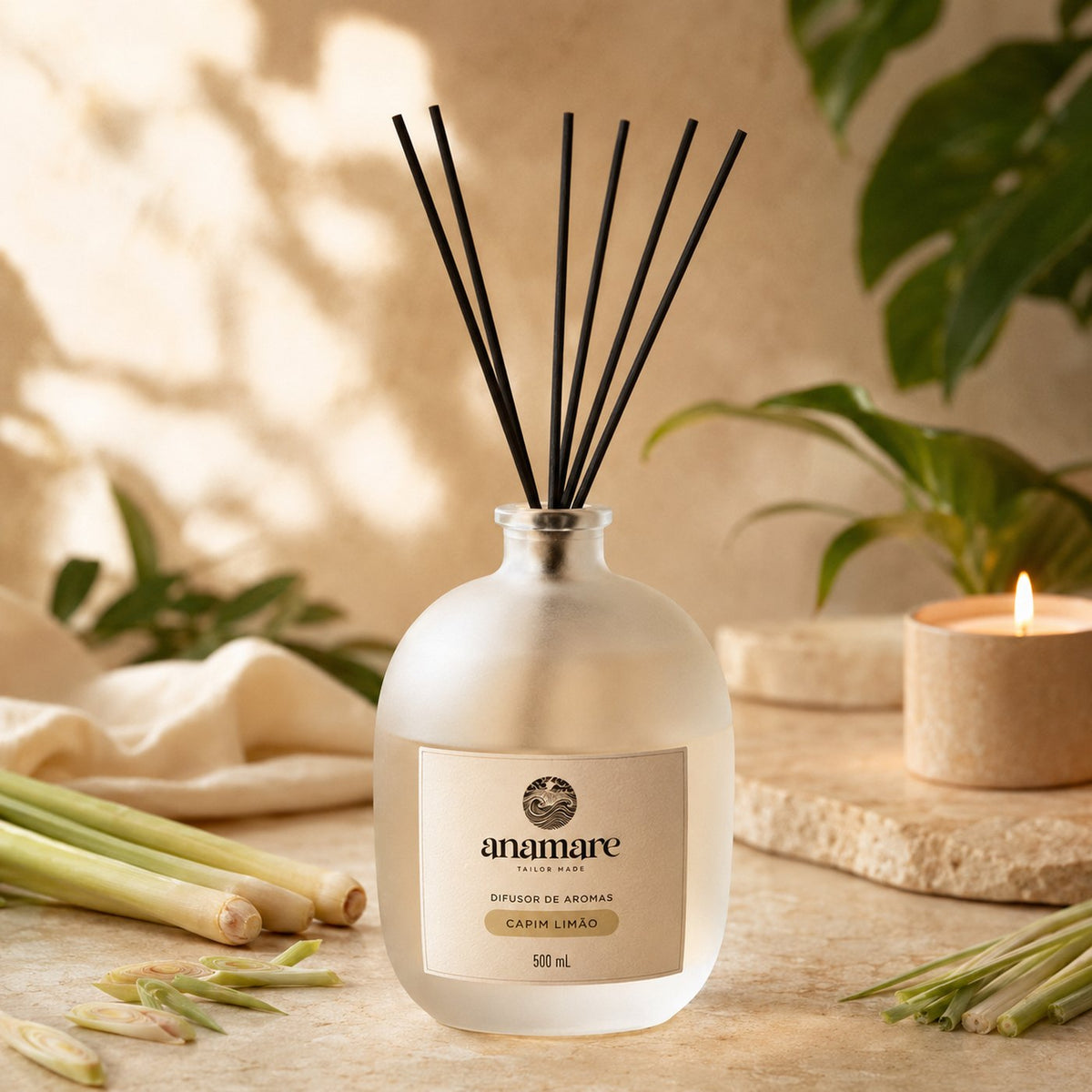 Difusor de Aromas Anamare Capim Limão de 500 mL, frasco de vidro fosco de ombro alto com gargalo prateado e varetas pretas, em superfície de pedra clara, com talos de capim-limão frescos e folhagem, em luz difusa