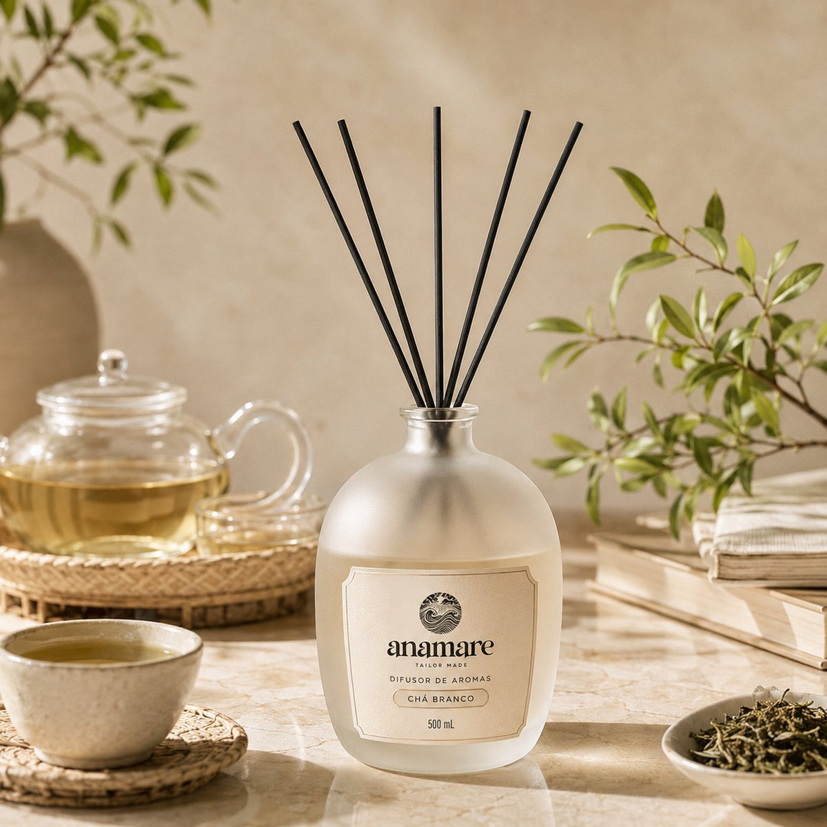 Difusor de Aromas Anamare Chá Branco de 500 mL, frasco de vidro fosco de ombro alto com gargalo prateado e varetas pretas, em bancada de mármore, com bule de vidro, xícara de chá, folhas secas e galho verde, em luz suave