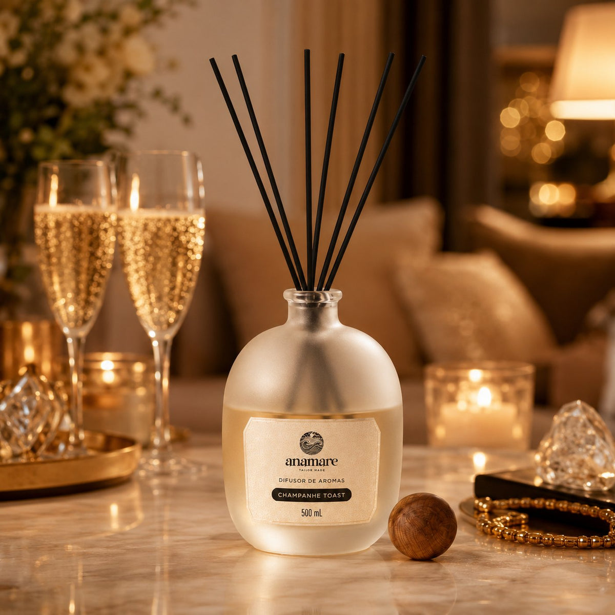 Difusor de Aromas Anamare Champanhe Toast de 500 mL, frasco de vidro fosco de ombro alto com gargalo prateado e varetas pretas, em mármore claro de sala em luz dourada, com duas taças de espumante, velas acesas, bandeja dourada e tampa esférica de madeira ao lado