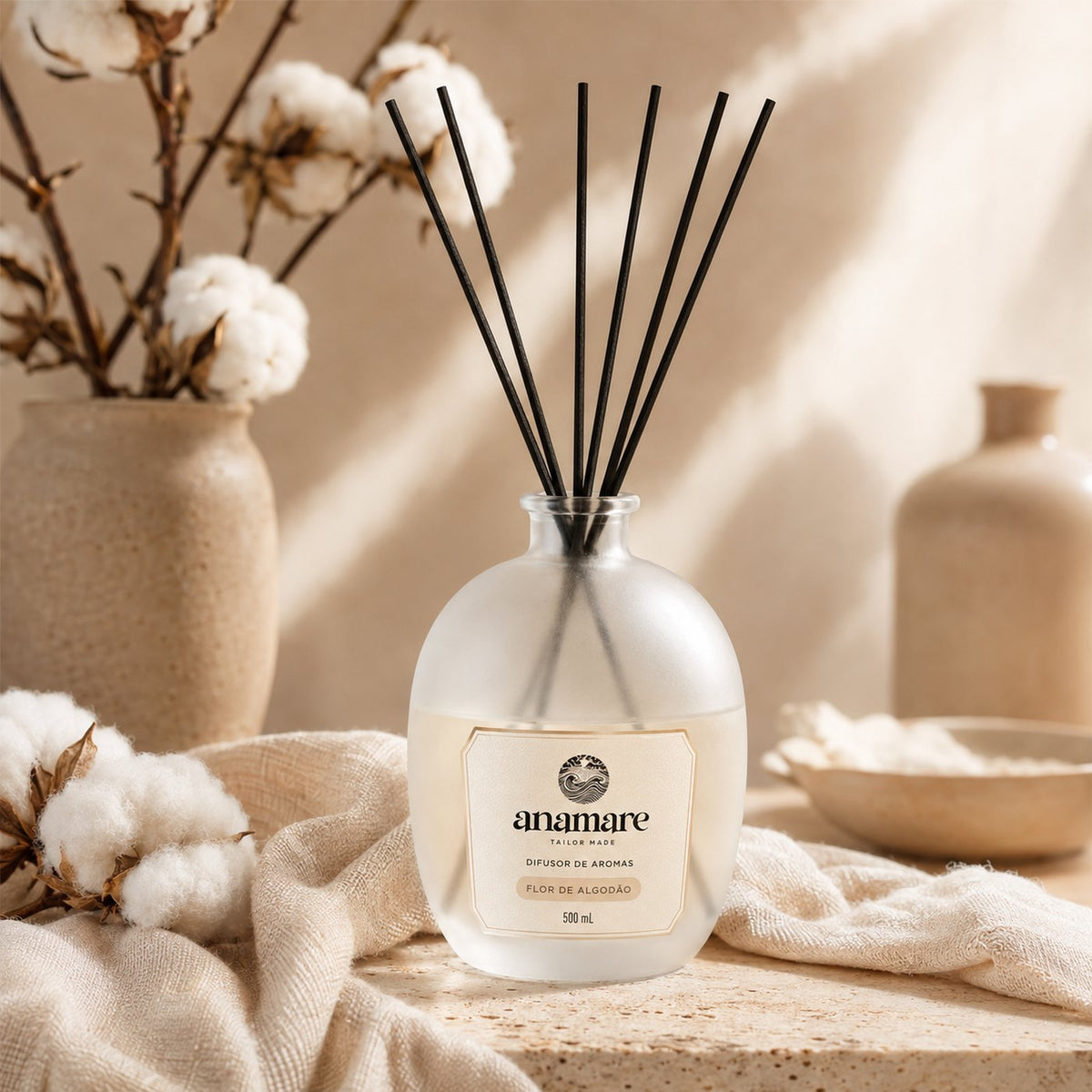 Difusor de Aromas Anamare Flor de Algodão de 500 mL, frasco de vidro fosco de ombro alto com gargalo prateado e varetas pretas, em pedra travertino, com galhos de algodão em vaso de cerâmica e tecido cru amassado, em luz lateral quente
