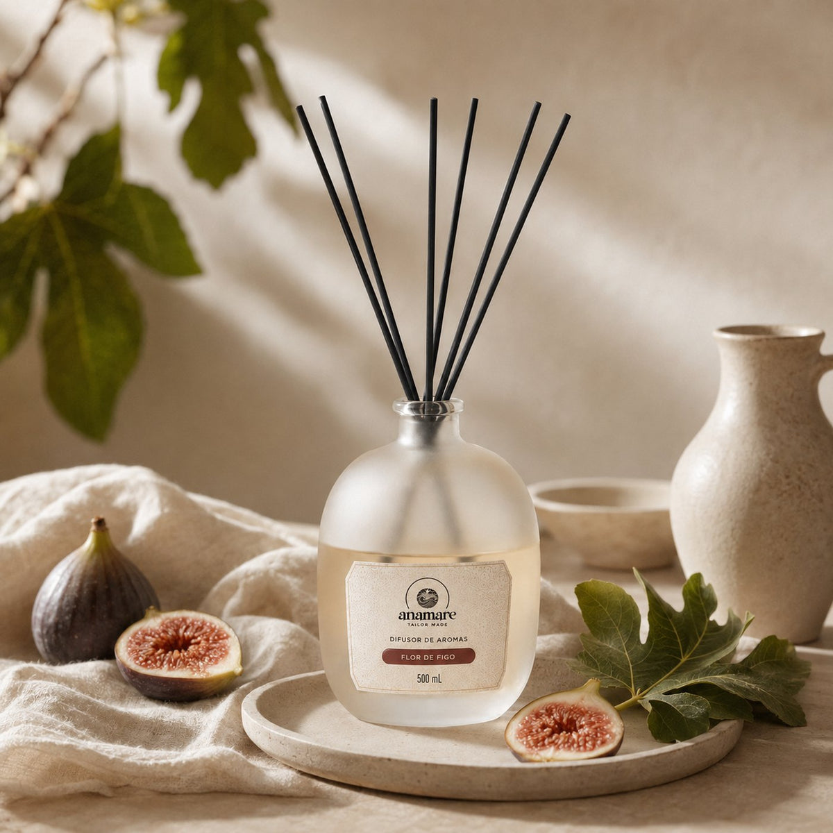Difusor de Aromas Anamare Flor de Figo de 500 mL, frasco de vidro fosco de ombro alto com gargalo prateado e varetas pretas, em prato de cerâmica sobre linho cru, com figos frescos partidos, folhas de figueira e jarra de cerâmica, em luz suave