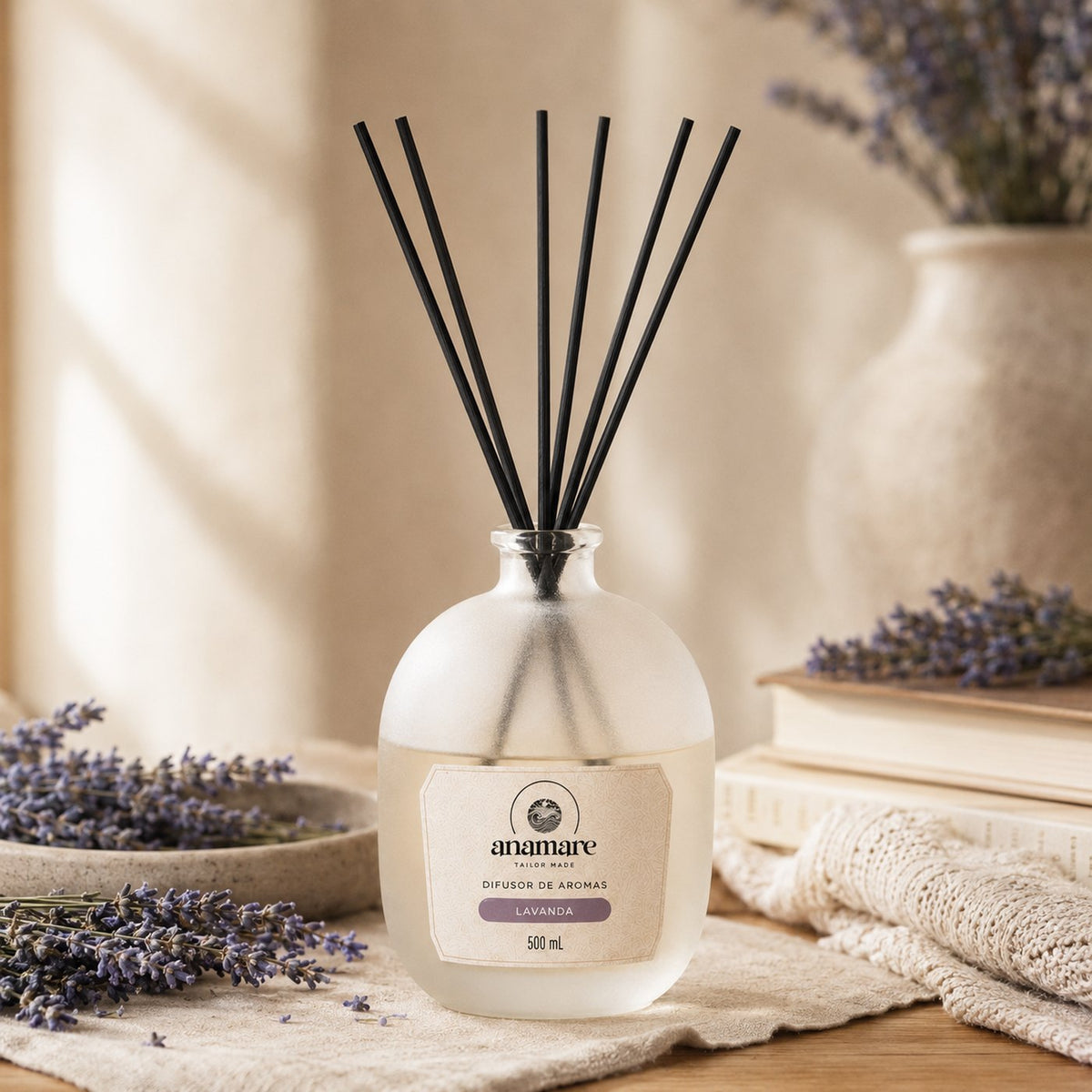 Difusor de Aromas Anamare Lavanda de 500 mL, frasco de vidro fosco de ombro alto com gargalo prateado e varetas pretas, em toalha de linho cru, com ramos de lavanda em prato de cerâmica, livros empilhados e manta de tricô, em luz de janela