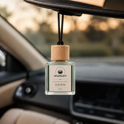 Difusor de Carro Anamare Alecrim, frasco de vidro quadrado de 10 mL com tampa de madeira e cordão preto, pendurado no retrovisor de um carro, com o painel desfocado e luz de fim de tarde pela janela