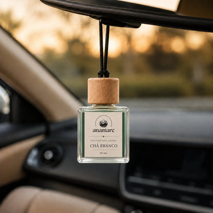 Difusor de Carro Anamare Chá Branco, frasco de vidro quadrado de 10 mL com tampa de madeira e cordão preto, pendurado no retrovisor de um carro, com o painel desfocado e luz de fim de tarde pela janela