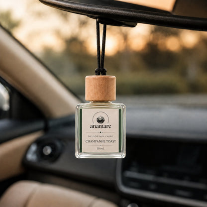 Difusor de Carro Anamare Champanhe Toast, frasco de vidro quadrado de 10 mL com tampa de madeira e cordão preto, pendurado no retrovisor de um carro, com o painel desfocado e luz de fim de tarde pela janela