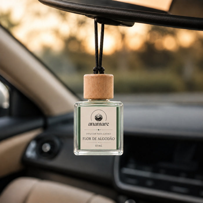 Difusor de Carro Anamare Flor de Algodão, frasco de vidro quadrado de 10 mL com tampa de madeira e cordão preto, pendurado no retrovisor de um carro, com o painel desfocado e luz de fim de tarde pela janela