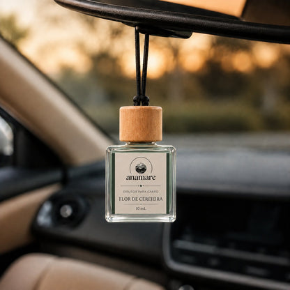 Difusor de Carro Anamare Flor de Cerejeira, frasco de vidro quadrado de 10 mL com tampa de madeira e cordão preto, pendurado no retrovisor de um carro, com o painel desfocado e luz de fim de tarde pela janela
