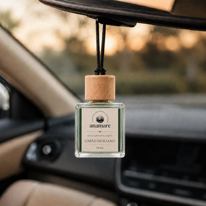 Difusor de Carro Anamare Limão Siciliano, frasco de vidro quadrado de 10 mL com tampa de madeira e cordão preto, pendurado no retrovisor de um carro, com o painel desfocado e luz de fim de tarde pela janela