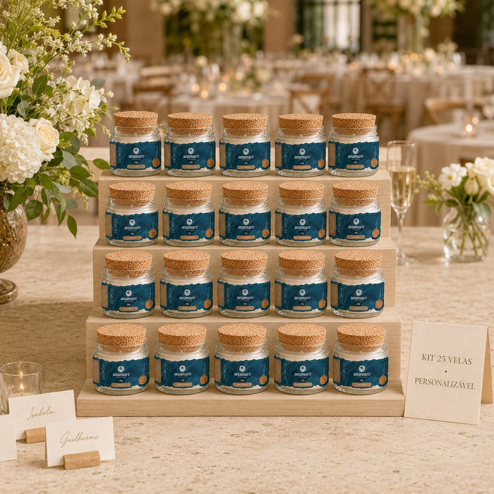 Kit de 25 lembrancinhas Anamare: velas Petit de 25 g em potes de vidro com tampa de cortiça e rótulo azul-petróleo personalizado, dispostas em escada de madeira clara sobre mesa de festa, ao lado de cartões de lugar com nomes dos noivos e arranjo de rosas brancas