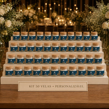 Kit de 50 lembrancinhas Anamare: velas Petit de 25 g em potes de vidro com tampa de cortiça e rótulo personalizado, em expositor de quatro degraus com placa gravada, diante do salão de festa iluminado