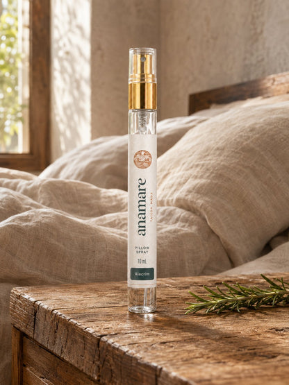 Pillow Spray Alecrim 10 mL, flaconete de vidro com válvula spray dourada, em mesa rústica de madeira ao sol, com ramo de alecrim fresco ao lado e roupa de cama de linho cru