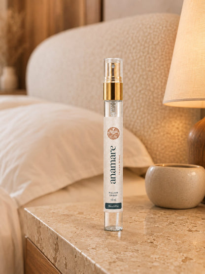 Pillow Spray Baunilha 10 mL, flaconete de vidro com válvula spray dourada, em mesa de travertino ao lado de abajur aceso, com poltrona de bouclé e xícara de cerâmica na cena
