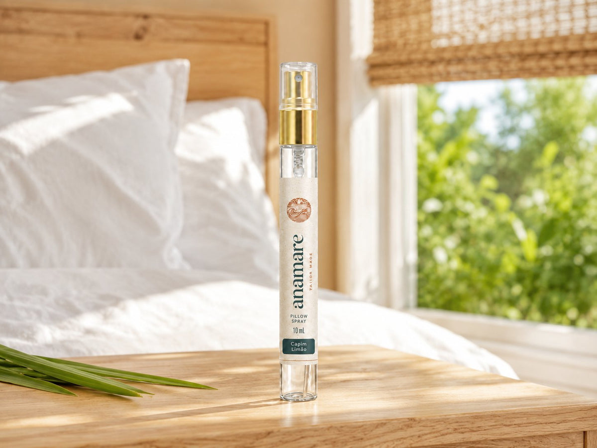Pillow Spray Capim Limão 10 mL, flaconete de vidro com válvula spray dourada, em mesa de madeira clara junto à janela aberta, com folhas de capim-limão ao lado e cama branca ao fundo